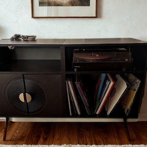 Puede incluir: Consola de tocadiscos negra de estilo moderno de mediados de siglo con tocadiscos, almacenamiento para discos de vinilo y una puerta de armario con dise&ntilde;o de disco de vinilo. La consola tiene una base blanca y patas anguladas.