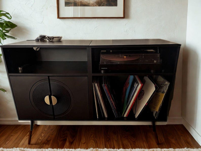 Puede incluir: Consola de tocadiscos negra de estilo moderno de mediados de siglo con tocadiscos, almacenamiento para discos de vinilo y una puerta de armario con dise&ntilde;o de disco de vinilo. La consola tiene una base blanca y patas anguladas.