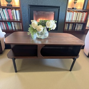 May include: A dark brown wooden coffee table with black legs. The table has a rectangular top with rounded corners and a lower shelf with an open circular design. A glass vase with white roses sits on top.