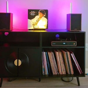 Puede incluir: Mueble de entretenimiento negro con dise&ntilde;o de disco de vinilo en las puertas del armario. La unidad tiene un tocadiscos, altavoces y una colecci&oacute;n de discos de vinilo. Un &aacute;lbum de Michael Jackson se muestra en la parte superior, iluminado por luces rosas y blancas.