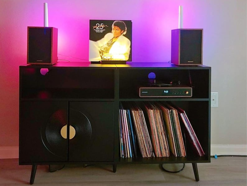 Puede incluir: Mueble de entretenimiento negro con dise&ntilde;o de disco de vinilo en las puertas del armario. La unidad tiene un tocadiscos, altavoces y una colecci&oacute;n de discos de vinilo. Un &aacute;lbum de Michael Jackson se muestra en la parte superior, iluminado por luces rosas y blancas.