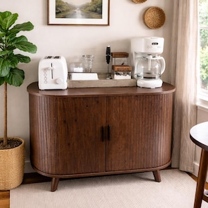 May include: A white toaster, coffee maker, and a wooden cabinet with a dark brown finish. The cabinet has two doors and is placed on a light-colored rug. A framed landscape painting and decorative woven wall hangings are also visible.