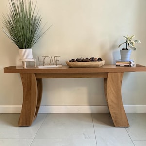 May include: A wooden console table with a curved leg design. The table is topped with decorative items, including a plant, a wooden bowl filled with pinecones, and a metal sign that reads "HOME".