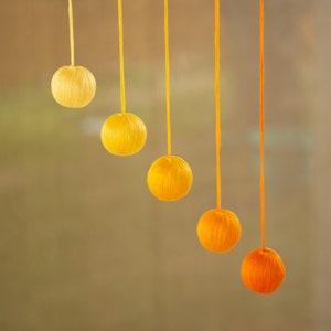May include: A wooden dowel rod with five yarn balls hanging from it. The yarn balls are different shades of orange and yellow, starting with a bright yellow at the top and transitioning to a darker orange at the bottom.