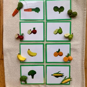 Puede incluir: Un conjunto de tarjetas con bordes verdes, cada una representando una fruta o verdura diferente. Cada tarjeta va acompañada de una pequeña réplica tridimensional del producto, incluyendo zanahorias, manzanas, plátanos y naranjas. Las tarjetas están dispuestas sobre una superficie blanca.