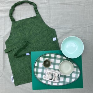 Puede incluir: Un delantal verde, un cuenco turquesa y una bandeja de servicio con un estampado de cuadros verdes y blancos. La bandeja contiene una taza pequeña, un cuenco pequeño y un cortador de huevos plateado. Los artículos están dispuestos sobre una superficie gris claro.