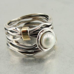 Puede incluir: Un anillo de plata con una perla y un acento dorado. El anillo está hecho de múltiples bandas que están entrelazadas.