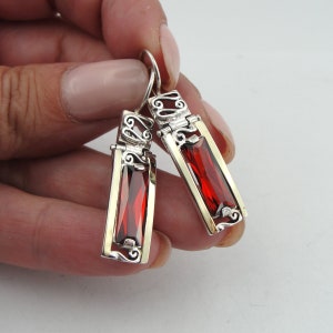 May include: Silver earrings with red gemstone accents. The earrings feature a delicate filigree design and dangle from a hook.