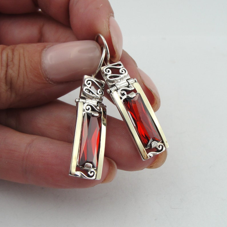 May include: Silver earrings with red gemstone accents. The earrings feature a delicate filigree design and dangle from a hook.