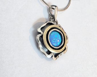 Puede incluir: Colgante de plata con una piedra central de ópalo azul, rodeada por un anillo dorado y un diseño floral plateado. El colgante cuelga de una cadena de plata. El ópalo tiene una cualidad iridiscente.