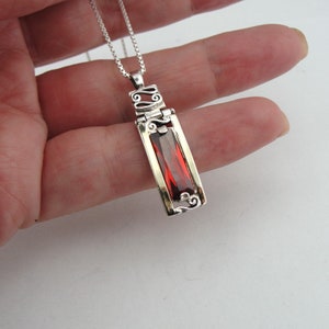 May include: A silver necklace with a red gemstone pendant. The pendant is rectangular and has a filigree design. The chain is a delicate box chain.