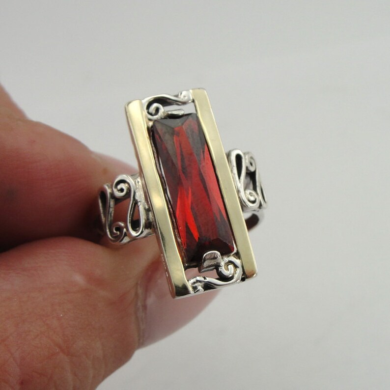 May include: A silver ring with a red gemstone set in a gold rectangle. The ring has a decorative design on the band.