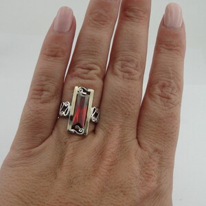 May include: A silver ring with a red gemstone set in a gold frame. The ring has a delicate, ornate design.