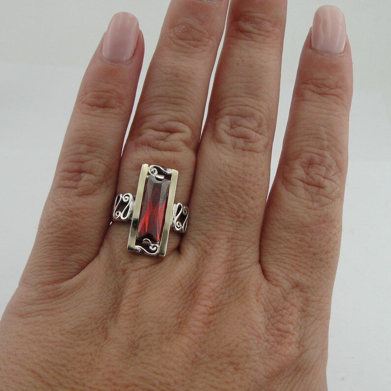 May include: A silver ring with a red gemstone set in a gold frame. The ring has a delicate, ornate design.