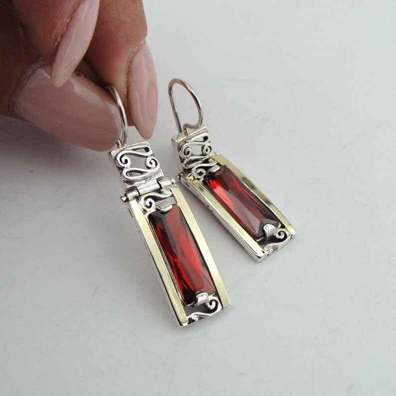 May include: A pair of silver dangle earrings with red gemstone accents. The earrings feature a delicate filigree design and a rectangular gemstone.