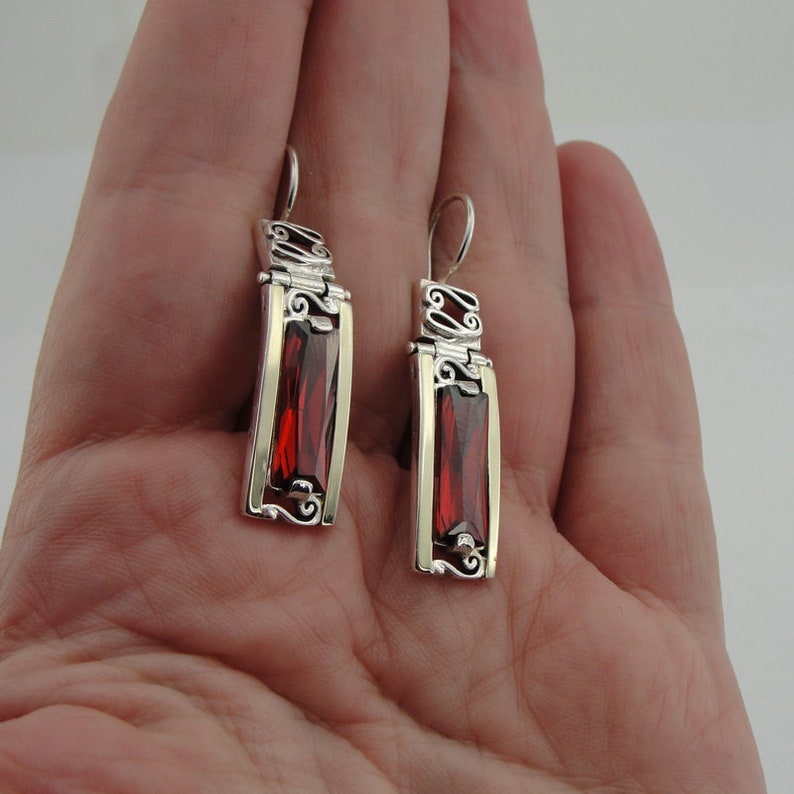 May include: A pair of silver dangle earrings with red gemstone accents. The earrings feature a delicate filigree design and a rectangular gemstone.