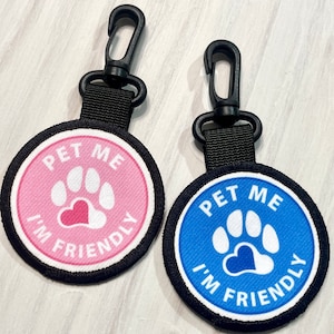 May include: Two round, black-edged, fabric patches with a white paw print and a heart in the center. The text "Pet Me I'm Friendly" is written around the paw print. The patches are attached to black clips.