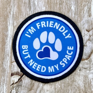 May include: Blue and white round patch with black stitching. It says "I'm Friendly But Need My Space" with a paw print in the center that has a blue heart in the middle.
