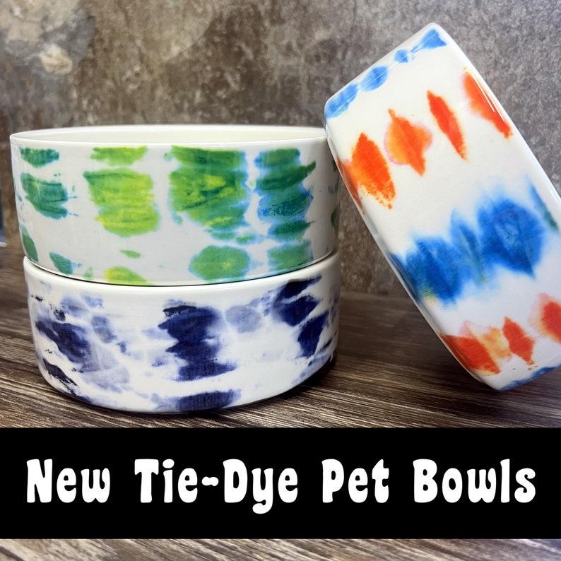May include: Three ceramic pet bowls with tie-dye patterns. The bowls are white with blue, green, and orange tie-dye designs.