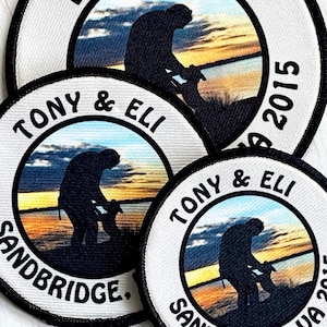 May include: Three round white patches with black borders. Each patch features a silhouette of a person with a dog in a sunset landscape. The text "Tony & Eli" and "Sandbridge" are printed on the patches. The year "2015" is printed on the top patch.