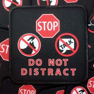 May include: A black patch with red and white symbols. The patch has a red stop sign, a red circle with a white dog and hand inside, a red circle with a white speech bubble with the word "TALK" inside, and the text "DO NOT DISTRACT" in red.