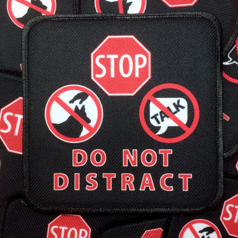 May include: A black patch with red and white symbols. The patch has a red stop sign, a red circle with a white dog and hand inside, a red circle with a white speech bubble with the word "TALK" inside, and the text "DO NOT DISTRACT" in red.