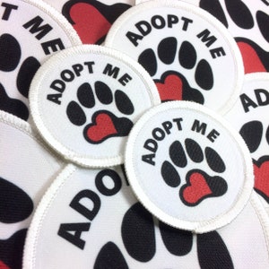 May include: A close-up of several white round patches with black paw prints and red hearts. The patches have the text "Adopt Me" printed on them.