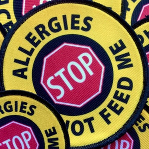 May include: Yellow round patch with black text that reads "ALLERGIES STOP NOT FEED ME". The text is in a circle around a red stop sign with white text that reads "STOP".