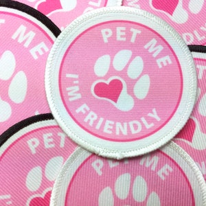 May include: Pink circular patch with the text "PET ME I'M FRIENDLY" around a white paw print with a pink heart in the center. The patch has a white border and is made of fabric.