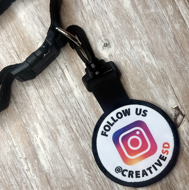 May include: Black and white round badge with the text "FOLLOW US" and the Instagram logo with the text "@CREATIVESD". The badge is attached to a black lanyard with a silver clip.