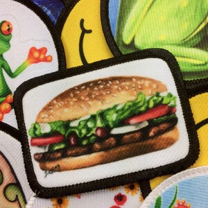 May include: A close-up of a white patch with a black border, featuring a detailed image of a hamburger with lettuce, tomato, and a sesame seed bun.
