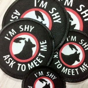 May include: A set of black and red round patches with the text "I'm Shy Ask to Meet Me" and a silhouette of a dog's head with a hand reaching out to pet it.