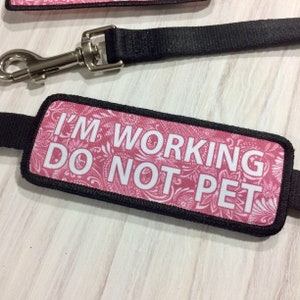 May include: A pink and white dog leash with a black strap and a black and white patch that says "I'm Working Do Not Pet".