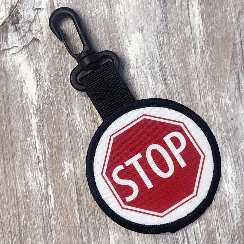 May include: A black keychain with a round, white and black fabric patch featuring a red stop sign with white lettering. The sign reads "STOP".