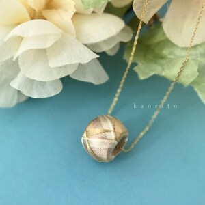 May include: A necklace featuring a spherical pendant wrapped in multi-toned beige and metallic gold thread, suspended from a thin gold-tone chain. The text "kaorito" is visible in small white letters near the pendant on the blue surface.