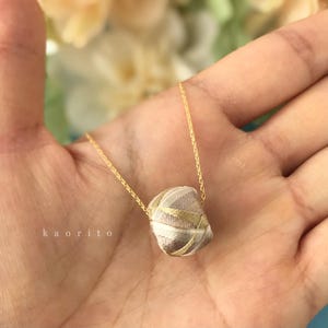 May include: A gold chain necklace featuring a small, rounded pendant wrapped in beige and metallic gold thread with a geometric pattern. The pendant is suspended from a delicate gold link chain. The text kaorito is visible on the left.