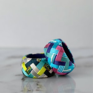 Könnte beinhalten: Zwei handgefertigte Stoffringe mit gewebtem Design. Ein Ring hat Türkis, Gelb und Silber. Der andere Ring zeigt eine Mischung aus Blau, Rosa und Türkis. Die Ringe sind wahrscheinlich Serviettenringe oder dekorative Akzente. Durchmesser ca. 5 cm.
