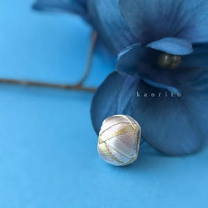 May include: A small, spherical bead wrapped in metallic gold and soft pink thread, featuring a textured, crisscross pattern. The bead is positioned in front of a dark blue flower petal, with the text kaorito visible in white lettering.