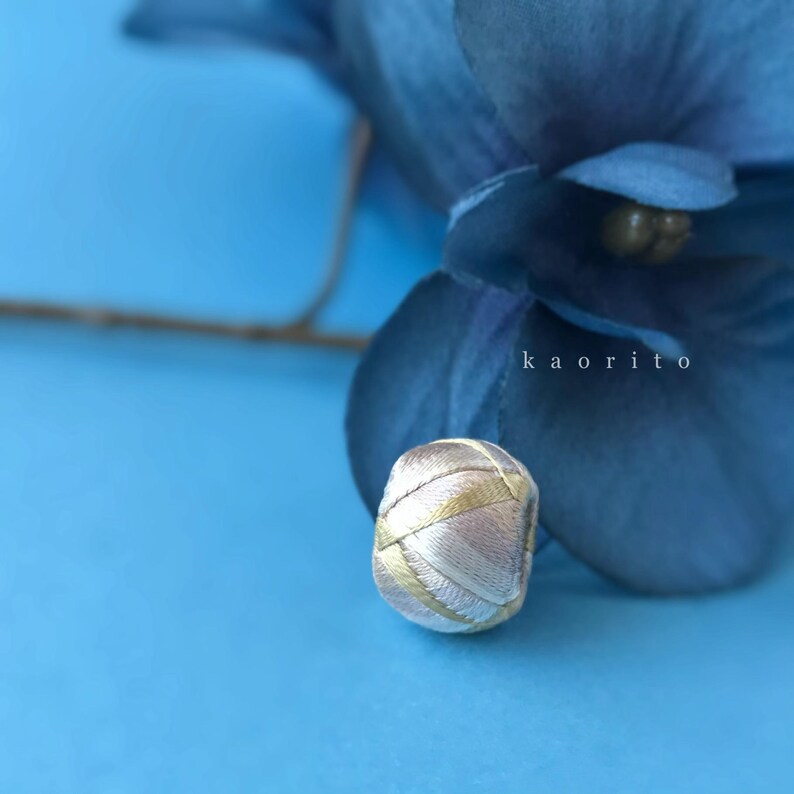 May include: A small, spherical bead wrapped in metallic gold and soft pink thread, featuring a textured, crisscross pattern. The bead is positioned in front of a dark blue flower petal, with the text kaorito visible in white lettering.