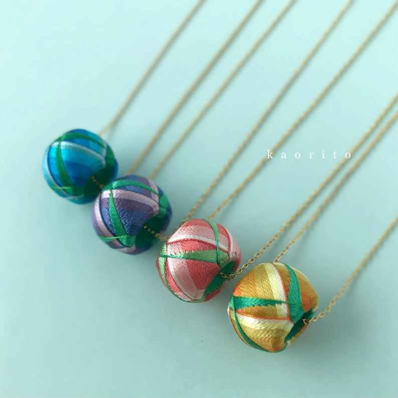 May include: Four necklaces featuring spherical pendants wrapped in multicolored silk thread with a geometric pattern. Each pendant is suspended on a thin gold-tone chain. The text kaorito is visible in the upper right area of the image.