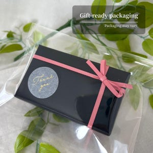 May include: A rectangular black gift box wrapped with a thin pink ribbon tied in a bow. A circular gray sticker with the gold cursive text Thank you is affixed to the top. The box is enclosed in a clear plastic sleeve for protection and presentation.