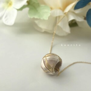 May include: A necklace featuring a spherical pendant wrapped in beige and metallic gold thread, suspended from a thin gold-tone chain. The text kaorito is printed in white on the right side of the pendant. The design is intricate and textured.