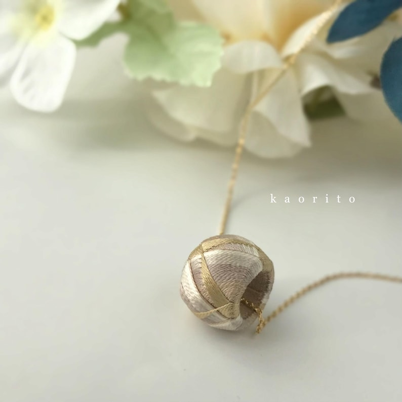 May include: A necklace featuring a spherical pendant wrapped in beige and metallic gold thread, suspended from a thin gold-tone chain. The text kaorito is printed in white on the right side of the pendant. The design is intricate and textured.