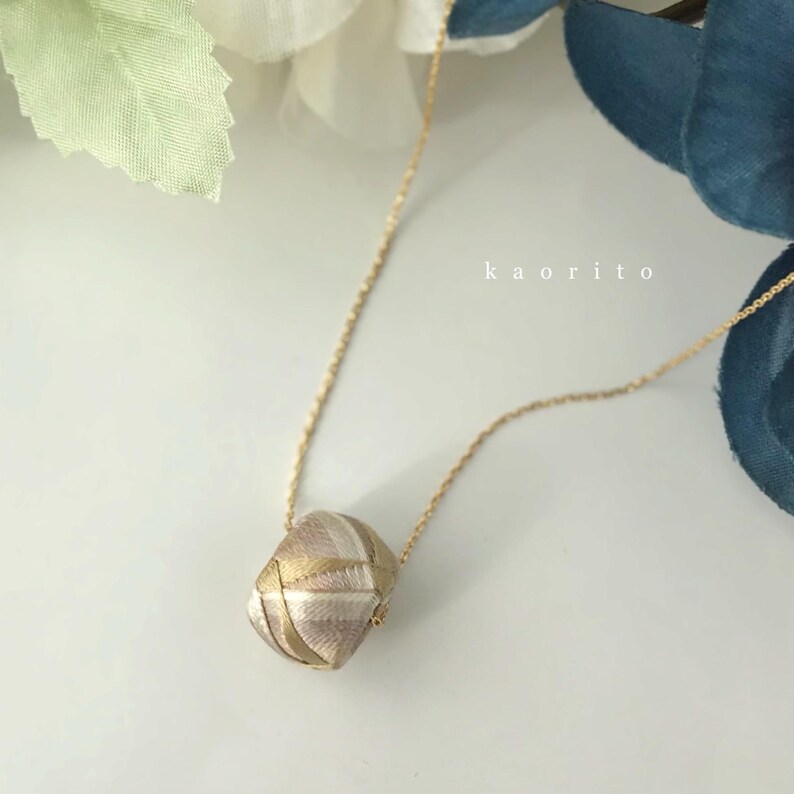 May include: A gold-tone chain necklace featuring a geometric, multi-faceted pendant wrapped in metallic gold and silver thread. The pendant displays a complex, woven pattern of intersecting lines. The text kaorito is visible in the background.