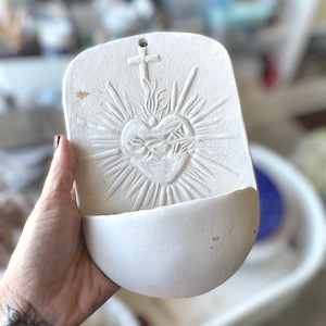 May include: A white ceramic wall pocket with a religious design. The pocket features a heart with flames, a cross, and sun rays. The bottom of the pocket is curved, and there is a hole at the top for hanging.