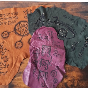 May include: Three pieces of leather with black ink symbols and designs. The leather is brown, green, and purple.