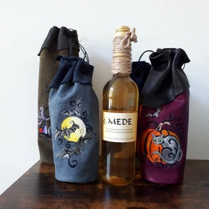 May include: A collection of wine bottle covers and a bottle of mead. The covers are in various colors, including dark green, blue, and purple, with embroidered Halloween-themed designs. The mead bottle has a rope-wrapped neck and a label that reads "MEDE".