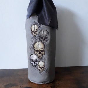 May include: Gray bottle sleeve with embroidered skulls and gears. The top is black and cinched with a drawstring. The skulls are in shades of white, gold, and gray.