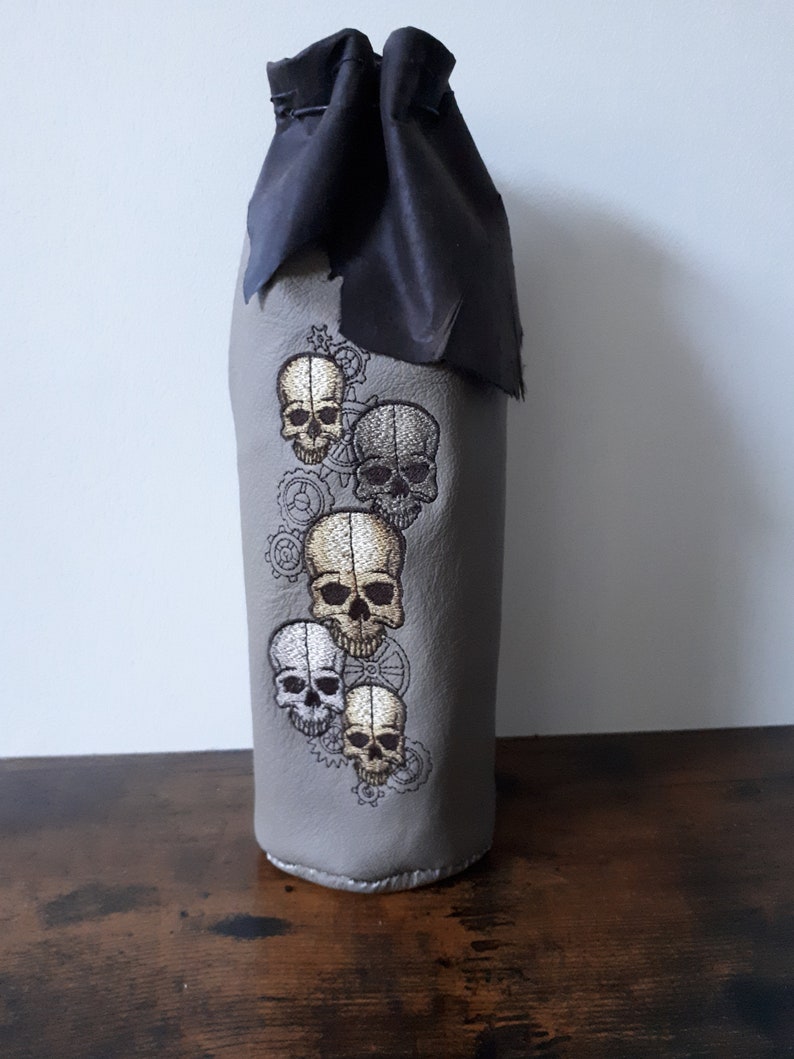 May include: Gray bottle sleeve with embroidered skulls and gears. The top is black and cinched with a drawstring. The skulls are in shades of white, gold, and gray.