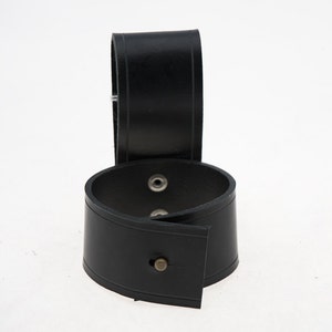 Puede incluir: Dos brazaletes de cuero negro con broches de metal plateado. Los brazaletes están apilados uno encima del otro.